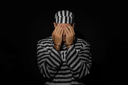 Remorseful prisoner in striped uniform with handcuffs hiding his face on black backgroundの写真素材