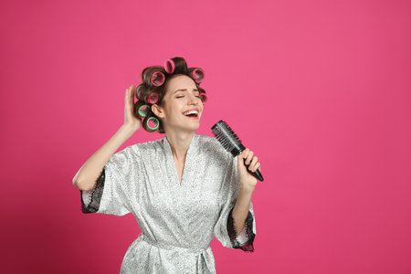 Beautiful young woman in silk bathrobe with hair curlers singing into hairbrush on pink background. Space for textの写真素材