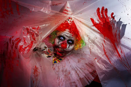 Terrifying clown staring through hole in torn bloodstained plastic film. Halloween party costumeの写真素材