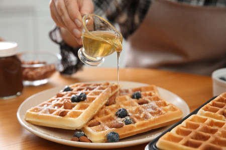 Woman pouring honey onto delicious Belgian waffles at wooden table in kitchen, closeup