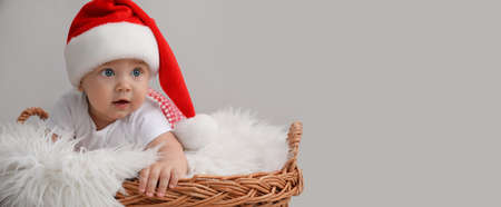 Cute baby wearing Santa hat in wicker basket on light gray background, banner design with space for text. Christmas celebrationの写真素材