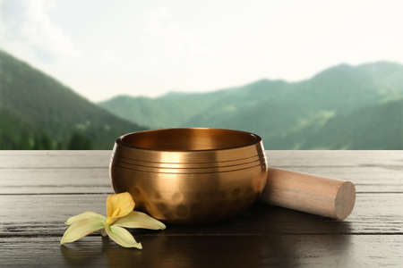 Golden singing bowl, mallet and flower on wooden table against mountain landscapeの写真素材