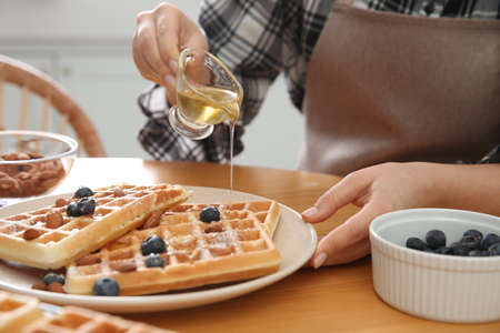 Woman pouring honey onto delicious Belgian waffles at wooden table in kitchen, closeupの写真素材