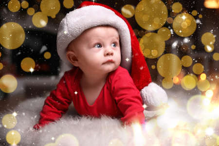 Cute little baby in red pajamas and Santa hat on floor against blurred festive lights. christmas suitの写真素材