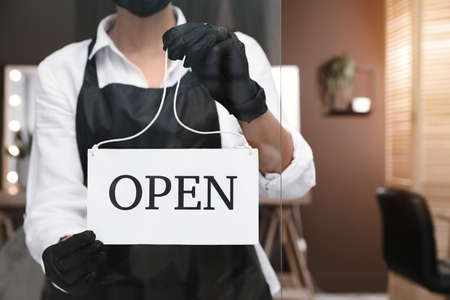 Woman hanging Open sign onto glass door in salon, closeup. Beauty services during quarantineの写真素材