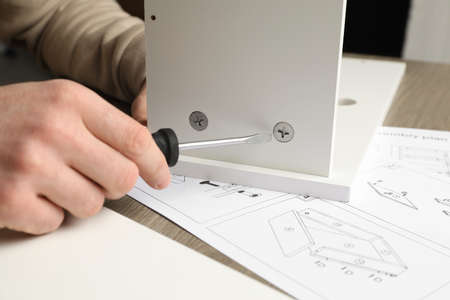 Man with screwdriver assembling white furniture at table, closeup