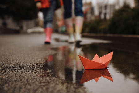 Little girl with her mother walking outdoors, focus on paper boat in puddle