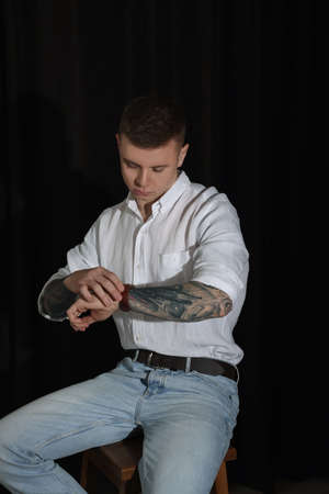 Young man with tattoos and wristwatch sitting on stool against dark background