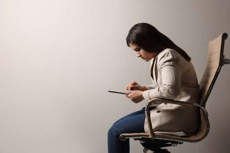 Young woman with bad posture using tablet while sitting on chair against gray background. Space for textの写真素材