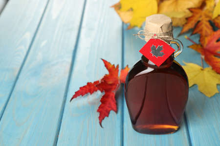 Glass bottle of tasty maple syrup and dry leaves on light blue wooden table. Space for textの写真素材