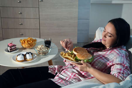 Happy overweight woman eating french fries with burgers at homeの写真素材