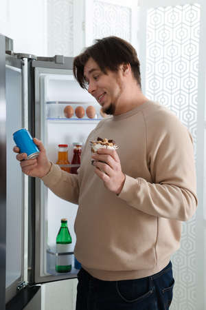 Overweight man holding dessert and tin can with beverage near open refrigerator in kitchenの写真素材