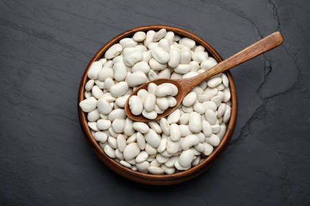 Raw white beans and spoon in bowl on black table, top view
