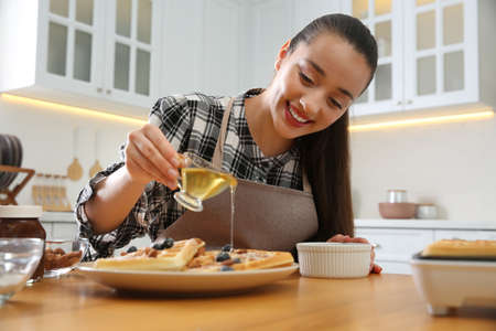 Woman pouring honey onto delicious Belgian waffles at wooden table in kitchen