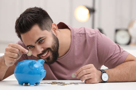 Happy young man putting money into piggy bank at white table indoorsの写真素材