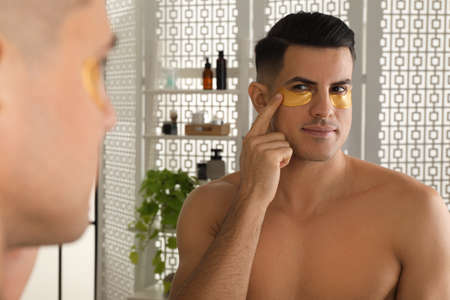 Man applying golden under eye patch near mirror at homeの写真素材