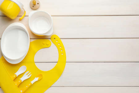 Flat lay composition with silicone baby bib and plastic dishware on white wooden background. Space for textの写真素材