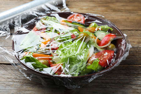 Bowl of fresh salad with plastic food wrap on wooden table, closeupの写真素材