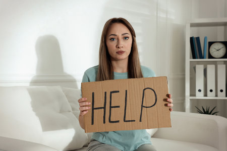 Unhappy young woman with HELP sign on sofa indoorsの写真素材