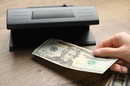Woman checking dollar banknote with currency detector at wooden table, closeup. money examination...