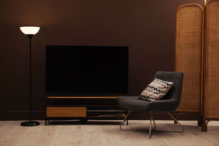 Modern TV on cabinet, armchair and floor lamp near brown wall in room. interior designの写真素材