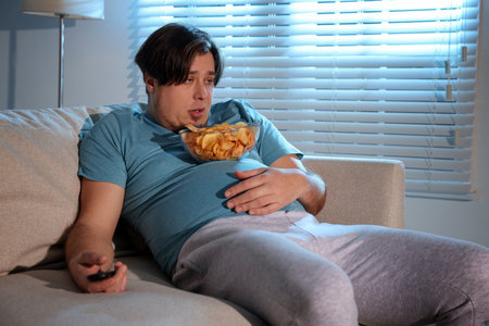 Overweight man with chips watching TV on sofa at homeの写真素材