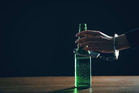 alcohol addiction. Woman handcuffed to bottle of beer against black background, closeup. Space for textの写真素材