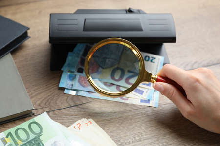 Woman checking Euro banknotes with currency detector and magnifying glass at wooden table, closeup....