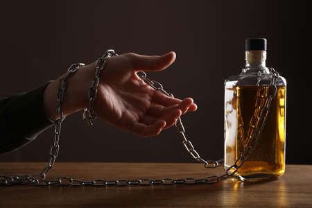 alcohol addiction. Man chained with bottle of whiskey at wooden table, closeupの写真素材