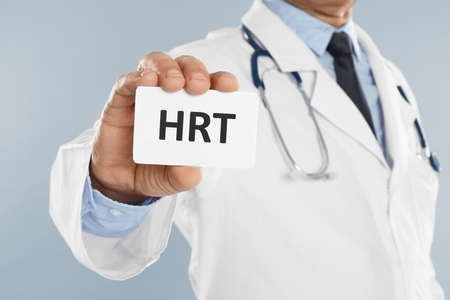 Doctor holding card with abbreviation HRT on light gray background, closeup. Hormone Replacement...