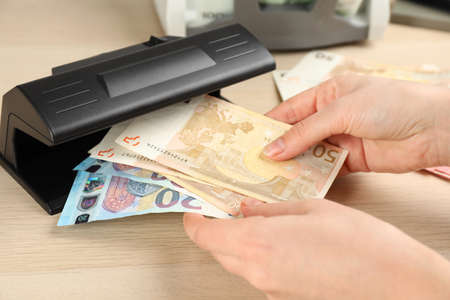 Woman checking Euro banknotes with currency detector at wooden table, closeup. Money examination...