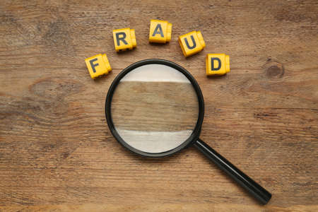 Word Fraud of yellow cubes with letters and magnifying glass on wooden background, flat layの写真素材