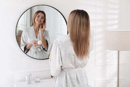 Beautiful young woman applying cream near mirror in roomの写真素材