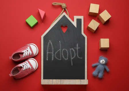 Small chalkboard with word ADOPT, toy bear, cubes and baby shoes on red background, flat layの写真素材