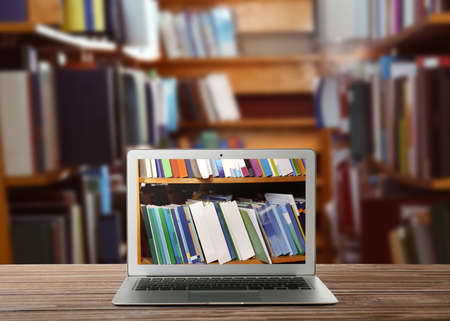 online library. Modern laptop on wooden table and shelves with books indoorsの写真素材