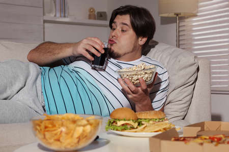Overweight man with bowl of popcorn and drink on sofa at homeの写真素材