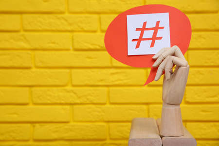 Wooden mannequin hand holding paper speech bubble with hashtag symbol near yellow brick wall. Space for textの写真素材