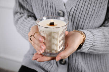 Woman with stylish jewelry holding burning soy candle on light background, closeup