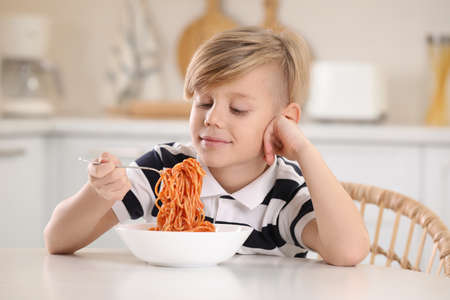 Happy boy eating tasty pasta at table in kitchenの写真素材