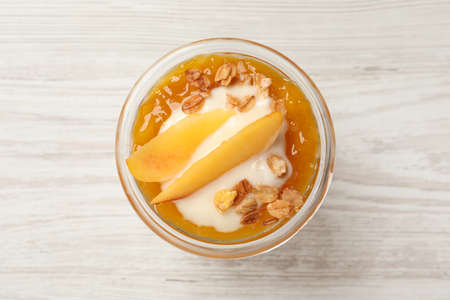 Tasty peach yogurt with granola, jam and pieces of fruit in glass on white wooden table, top viewの写真素材