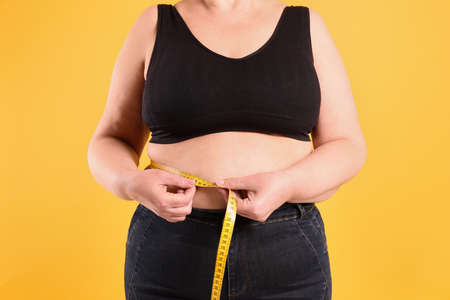 Overweight woman measuring waist with tape on yellow background, closeupの写真素材
