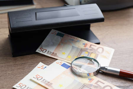 Modern currency detector with Euro banknotes and magnifying glass on wooden table. money examination...