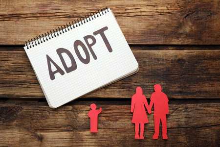 Figures of parents with child and word Adopt in notebook on wooden table, flat layの写真素材