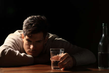 Addicted man with alcoholic drink at wooden table against black background, focus on glassの写真素材