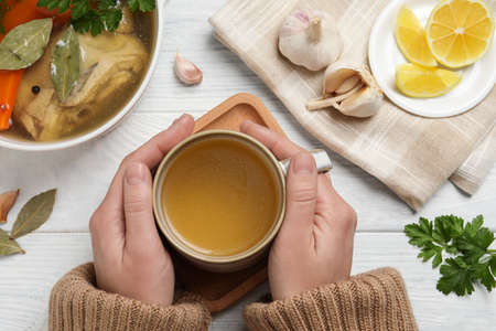 Woman with cup of hot delicious bouillon on white wooden table, top view