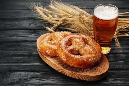Tasty pretzels, glass of beer and wheat spikes on black wooden table