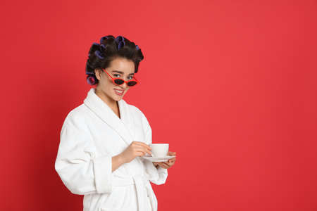 Happy young woman in bathrobe with hair curlers and sunglasses holding cup of drink on red background, space for textの写真素材