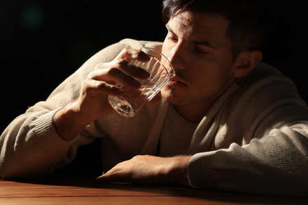Addicted man drinking alcohol on wooden table against black backgroundの写真素材