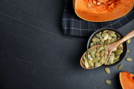 Bowl with seeds, wooden spoon and cut pumpkin on black table, flat lay. Space for textの写真素材