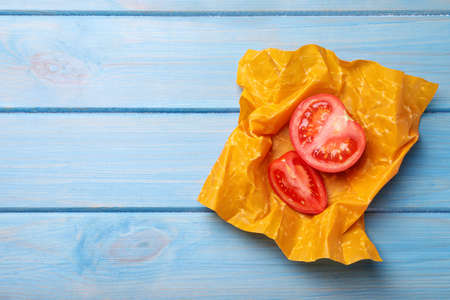 Slices of fresh tomato in beeswax food wrap on light blue wooden table, top view. Space for text
